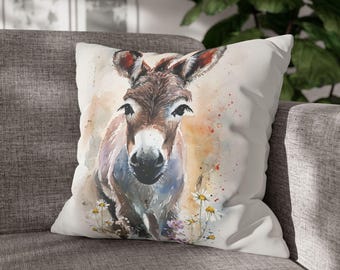 May include: A decorative throw pillow featuring a watercolor painting of a donkey. The donkey is brown and white with large ears and is surrounded by wildflowers. The pillow is on a gray couch.
