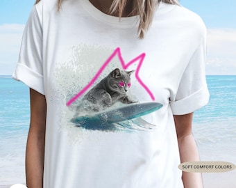 Puede incluir: Camiseta blanca con un gato gris surfeando sobre una tabla de surf azul, con un contorno de montaña rosa. El gato tiene ojos rosas. La camiseta tiene la frase "SOFT COMFORT COLORS".