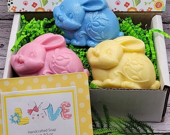 May include: A gift box filled with three bunny-shaped soaps. The soaps are blue, pink, and yellow. The box is lined with green grass and has a yellow and white polka dot label that says "Handcrafted Soap Net wt. 9.5 oz".