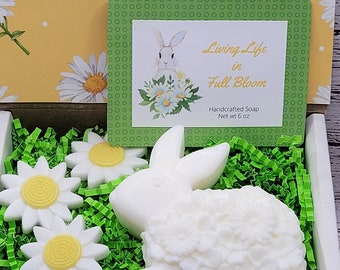 May include: A white bunny-shaped soap bar with a floral design, nestled in green shredded paper. The soap is in a white box with a green label that reads "Living Life in Full Bloom" and "Handcrafted Soap Net wt 6 oz".