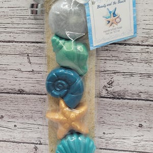 May include: A clear cellophane bag contains five seashell-shaped soaps in shades of silver, teal, blue, and gold, resting on sand. A tag reads "Beauty and the Beach" with a starfish graphic. The soaps are tied with a silver ribbon.