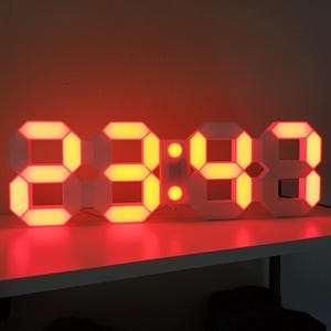 Op de afbeelding: Een witte digitale wandklok met een rechthoekig frame en grote, oplichtende rode LED-cijfers die de tijd 23:48 weergeven. De klok heeft een modern ontwerp met gesegmenteerde cijferdisplays en twee ronde stippen als scheidingsteken.