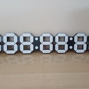May include: A modern digital clock with a black frame and white illuminated numbers. The clock displays the time in a segmented format, with each digit clearly visible. The clock is rectangular and rests on a wooden surface.