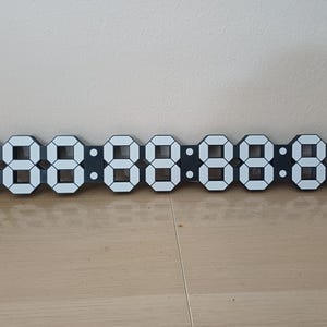 May include: A digital clock with a black frame and white LED numbers. The clock displays eight digits in a segmented format, showing the time. The clock is rectangular and sits on a wooden surface against a neutral wall.