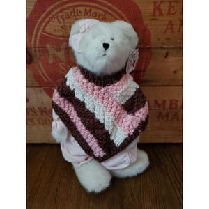 May include: A white teddy bear wearing a pink and brown striped poncho and a matching bow. The bear is also wearing a pink outfit. The background is a wooden crate with red lettering.