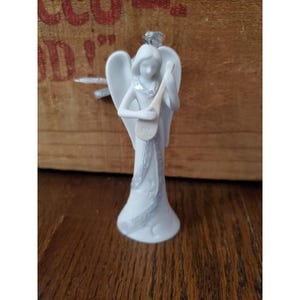 Op de afbeelding: Een witte keramische engelenfiguur met uitgespreide vleugels, die een snaarinstrument bespeelt. De engel draagt een lange jurk met decoratieve versieringen. Bovenop is een zilveren lint bevestigd om op te hangen. Het beeldje staat op een houten oppervlak.