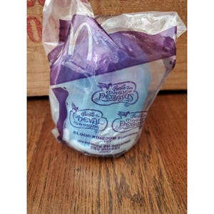 Puede incluir: Un juguete sellado de McDonald's de la serie Barbie Magic Pegasus. El embalaje es de plástico transparente con detalles en morado y presenta el logotipo de Barbie y texto, incluyendo "Cloud Kingdom Surprise Toy". El juguete del interior parece ser azul claro.