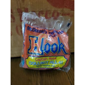 May include: Sealed McDonald's toy in a clear plastic bag. The bag features an orange and blue design with the word "Hook" in large letters. Text on the bag reads "Captain Hook COLLECT ALL 4!" and "McDonald's".