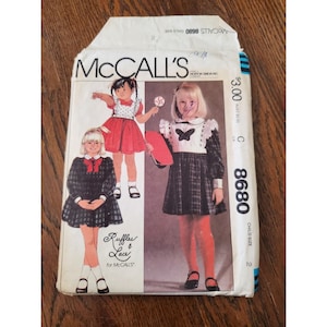May include: A McCall's sewing pattern for a child's dress. The pattern is for size 2 and includes two different styles. The first style is a red dress with a white bib and the second style is a plaid dress with a white bib and a butterfly applique. The pattern is titled "Ruffles & Lace for McCall's".