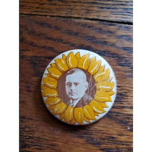 May include: A vintage button featuring a portrait of a man with glasses, surrounded by a yellow sunflower design.