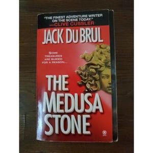 Może przedstawiać: Książka w miękkiej okładce zatytułowana "The Medusa Stone" autorstwa Jacka Du Brula. Okładka jest w przeważającej mierze czerwona z tytułem dużymi białymi literami. Z prawej strony znajduje się złota ilustracja twarzy kobiety. Książka zawiera cytat Clive'a Cusslera.