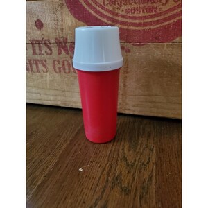 May include: A red plastic container with a white lid. The container is about 5 inches tall and has a diameter of about 2 inches.