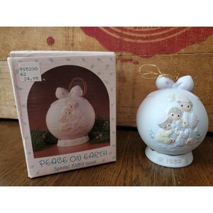 May include: A white ceramic Christmas ornament with a family scene, a bow, and a gold string for hanging. The ornament is from the 1989 "Peace on Earth" special issue. The ornament is in its original box, which also features the ornament design.