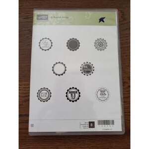 May include: A clear plastic case containing a set of eight rubber stamps. The stamps feature circular designs with various phrases like "You make me smile," "Be mine," and "For you." The set is titled "A Round Array."