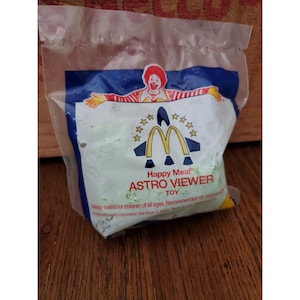 May include: A sealed Happy Meal toy package featuring the McDonald's logo and Ronald McDonald. The package reads "Happy Meal ASTRO VIEWER TOY". The package is clear plastic with a blue and white design and a green interior.