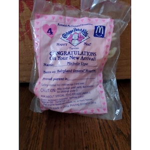 May include: Sealed Cabbage Patch Kids Happy Meal figurine in a pink plastic bag. The bag features the McDonald's logo, a heart with the number 4, and text that reads "CONGRATULATIONS On Your New Arrival!" The name "Michelie Elyse" is also printed on the bag.