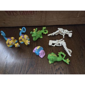 May include: A collection of colorful, small plastic toy figures, including two yellow and purple creatures, two green creatures, two white skeletal figures, and a blue, pink, and purple object. The toys are arranged on a wooden surface.