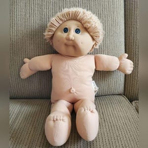 May include: A Cabbage Patch Kid doll with blonde hair and blue eyes. The doll is wearing a knitted hat and is sitting on a beige sofa.