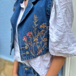 May include: A blue denim vest with embroidered flowers in shades of pink, brown, and green.