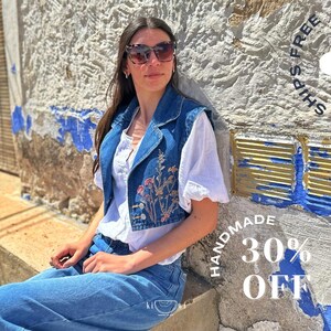 Puede incluir: Un chaleco vaquero con bordado floral y una blusa blanca. El chaleco tiene una parte delantera abotonada y un cuello. La imagen tambi&eacute;n muestra vaqueros azules y gafas de sol. El texto "HANDMADE 30% OFF" es visible.