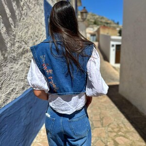 Puede incluir: Un chaleco vaquero con bordado floral, usado sobre una blusa blanca, combinado con vaqueros azules. El chaleco presenta un dise&ntilde;o corto y un cuello. El atuendo es casual y elegante, adecuado para el uso diario.