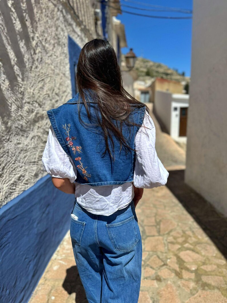 Puede incluir: Un chaleco vaquero con bordado floral, usado sobre una blusa blanca, combinado con vaqueros azules. El chaleco presenta un dise&ntilde;o corto y un cuello. El atuendo es casual y elegante, adecuado para el uso diario.