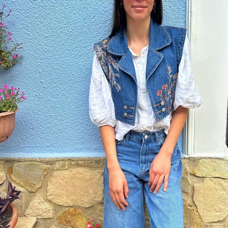 Puede incluir: Un chaleco vaquero azul con bordados florales en tonos rojos, naranjas y verdes. El chaleco tiene cuello y botones. La modelo lleva una blusa blanca y vaqueros azules.