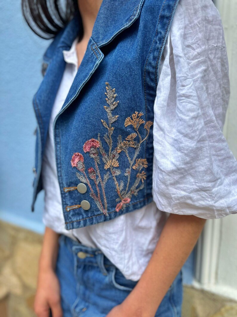 May include: A blue denim vest with embroidered flowers in shades of pink, brown, and green.