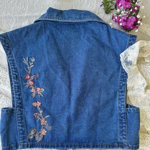 Puede incluir: Un chaleco vaquero sin mangas en un lavado azul medio. La parte trasera del chaleco presenta un dise&ntilde;o floral bordado en tonos rosa, melocot&oacute;n y verde. El chaleco tiene un cuello cl&aacute;sico y dos bolsillos delanteros.