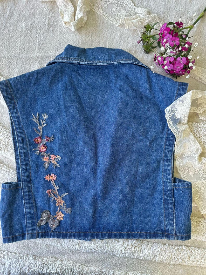 Puede incluir: Un chaleco vaquero sin mangas en un lavado azul medio. La parte trasera del chaleco presenta un dise&ntilde;o floral bordado en tonos rosa, melocot&oacute;n y verde. El chaleco tiene un cuello cl&aacute;sico y dos bolsillos delanteros.