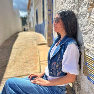 Puede incluir: Un chaleco vaquero azul con bordado floral, usado sobre una camiseta blanca, combinado con vaqueros azules. El chaleco presenta un dise&ntilde;o corto y cierre de botones. El atuendo es casual y elegante, adecuado para el uso diario.