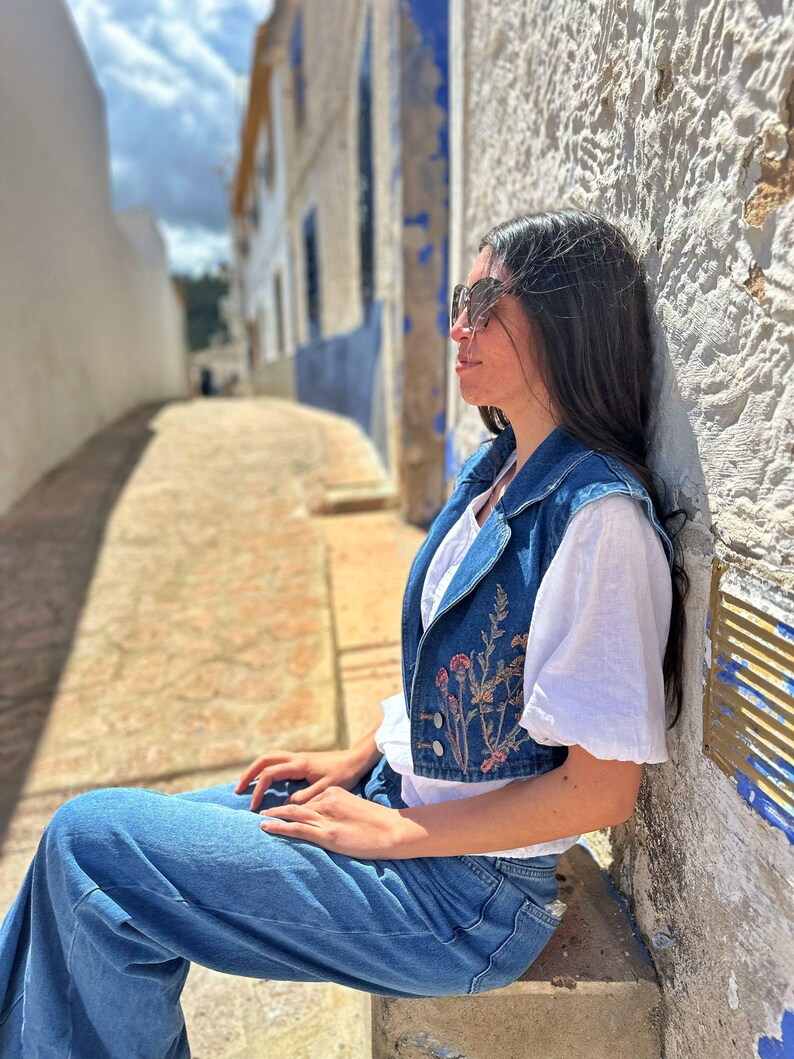 Puede incluir: Un chaleco vaquero azul con bordado floral, usado sobre una camiseta blanca, combinado con vaqueros azules. El chaleco presenta un dise&ntilde;o corto y cierre de botones. El atuendo es casual y elegante, adecuado para el uso diario.