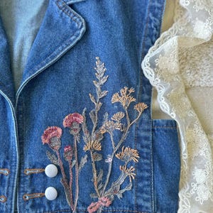 Puede incluir: Un chaleco vaquero sin mangas en un lavado azul medio, con detalles florales bordados en rosa, melocot&oacute;n y plata. El chaleco tiene cuello, dos botones blancos y un bolsillo. Un ribete de encaje blanco es visible en el lateral.