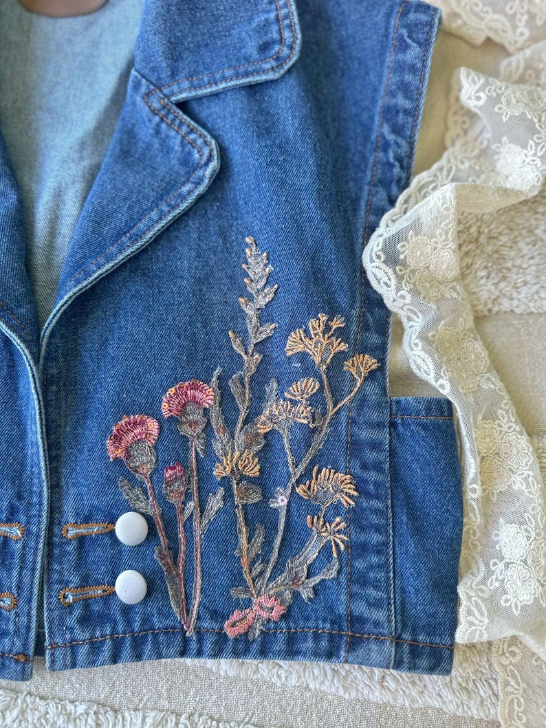 Puede incluir: Un chaleco vaquero sin mangas en un lavado azul medio, con detalles florales bordados en rosa, melocot&oacute;n y plata. El chaleco tiene cuello, dos botones blancos y un bolsillo. Un ribete de encaje blanco es visible en el lateral.