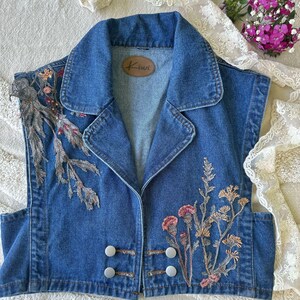 Puede incluir: Un chaleco vaquero sin mangas con cuello y dos filas de botones blancos. El chaleco presenta dise&ntilde;os florales bordados en tonos rosa, beige y verde. Una etiqueta de cuero marr&oacute;n con la palabra "Kiwi" est&aacute; cosida en el interior.