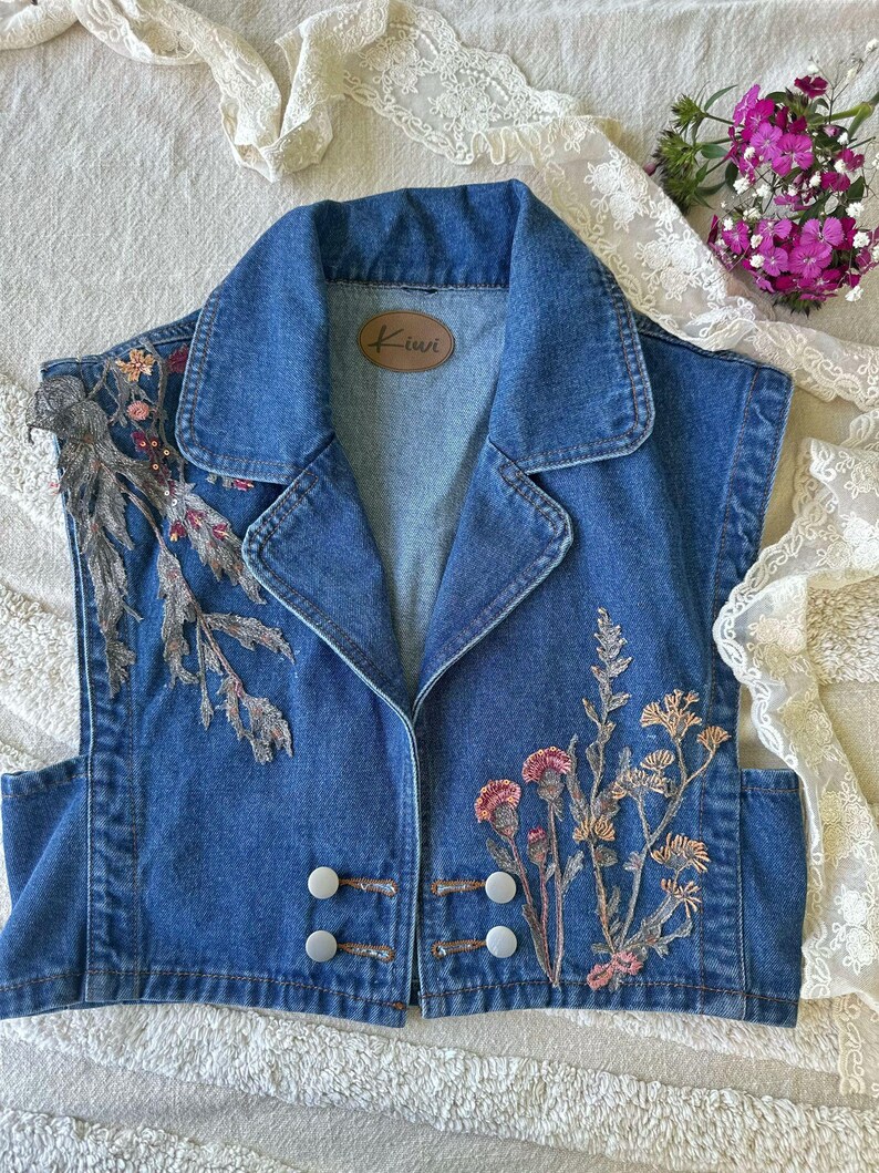 Puede incluir: Un chaleco vaquero sin mangas con cuello y dos filas de botones blancos. El chaleco presenta dise&ntilde;os florales bordados en tonos rosa, beige y verde. Una etiqueta de cuero marr&oacute;n con la palabra "Kiwi" est&aacute; cosida en el interior.