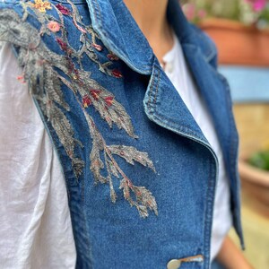 Puede incluir: Un chaleco vaquero azul con intrincados bordados florales en tonos grises, rosas y amarillos. El chaleco presenta un cuello cl&aacute;sico, dos botones decorativos y un dise&ntilde;o sin mangas. El bordado se concentra en la parte superior izquierda.