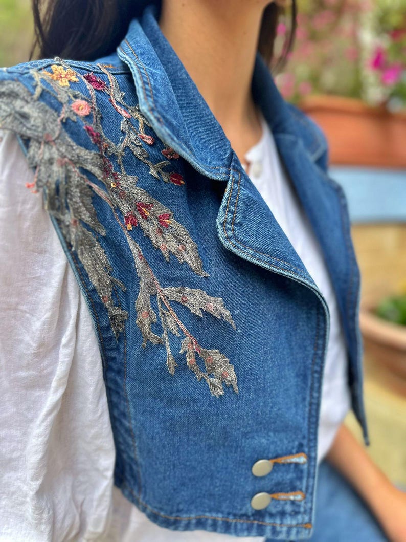 Puede incluir: Un chaleco vaquero azul con intrincados bordados florales en tonos grises, rosas y amarillos. El chaleco presenta un cuello cl&aacute;sico, dos botones decorativos y un dise&ntilde;o sin mangas. El bordado se concentra en la parte superior izquierda.