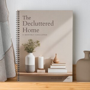 Op de afbeelding: Een spiraalgebonden boek met de titel "The Decluttered Home: A Monthly Challenge" op de omslag. De omslag heeft een beige achtergrond met een stilleven van een vaas met gedroogde bloemen, een witte kaars, een houten peer en een stapel boeken. Het boek staat op een houten plank.