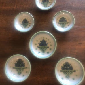 May include: Set of six small ceramic bowls. Each bowl features a white interior with a blue and green floral design. The rims are outlined in green. The bowls are arranged on a dark wooden surface.