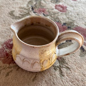 May include: A ceramic creamer pitcher featuring a light beige base with a speckled texture and a series of embossed arch patterns around the lower body. The piece has a wide handle and a distinct pouring spout with warm brown glaze accents.