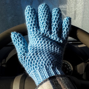 May include: A blue crocheted mitten with a textured pattern. The mitten is worn on a person's hand, which is holding a steering wheel.