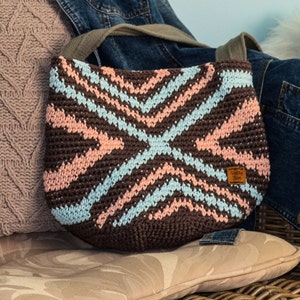 Könnte beinhalten: Eine braune gehäkelte Tasche mit einem geometrischen Muster in Rosa und Blau. Die Tasche hat ein braunes Leder-Etikett mit dem Text "Knotty Yarn" darauf.