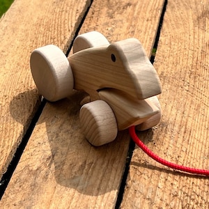 Pode incluir: Um brinquedo de madeira para puxar com um animal estilizado de cor castanho claro e uma corda vermelha presa na parte de trás.