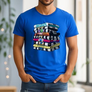 May include: A blue t-shirt featuring a graphic of a stack of colorful, folded shirts. The shirts in the graphic have various designs and text, including the word "ISELD". The t-shirt is worn by a person.