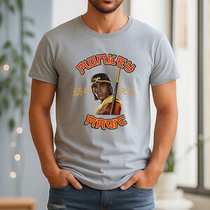 May include: A light gray t-shirt featuring a graphic of a person with a staff, with the words "MONKEY MAGIC" in red and gold. The shirt also has Chinese characters. The shirt is short-sleeved.