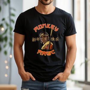 May include: Black t-shirt with a graphic featuring a portrait of a person with a staff, the words "MONKEY MAGIC" in red and gold, and Chinese characters. The shirt is short-sleeved.