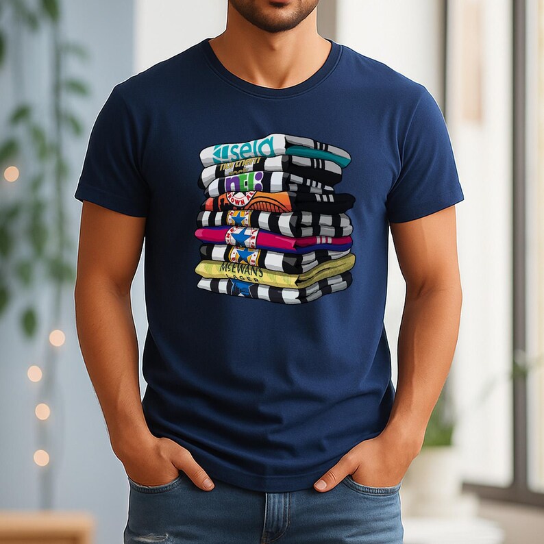 May include: Navy blue t-shirt featuring a graphic of a stack of folded shirts. Each shirt in the stack has a different color and design, with text visible on some. The shirts are stacked on top of each other.