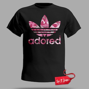 May include: Black t-shirt with a pink camouflage trefoil logo and the word "adored" in matching lettering. A red tag with "Love It Designs" is attached. The design is a modern take on a classic logo.
