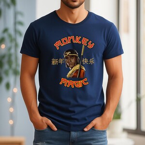 May include: Navy blue t-shirt featuring a graphic of a person with a staff, with the words "MONKEY MAGIC" in gold lettering. Chinese characters are also present. The shirt is a crew neck and made of a soft material.
